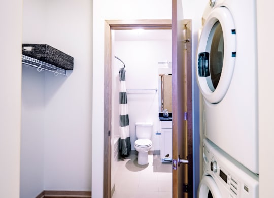 Laundry room here at 7west Apartment Homes with a stacked washer and dryer on the right, and a door opening into a bathroom with a toilet, sink, and striped shower curtain in neutral tones.