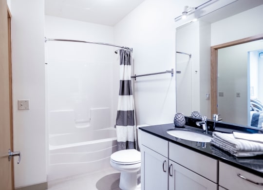 Bright, modern bathroom here at 7west Apartment Homes with white walls, a bathtub with a striped curtain, and a black countertop sink. Clean, minimalist design conveys tranquility.