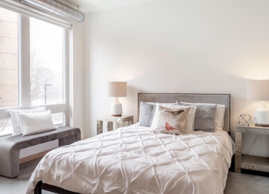 Bright bedroom here at 7west Apartment Homes with a neatly made bed, featuring white and gray linens and a decorative bird pillow. Soft lighting from two lamps creates a cozy ambiance.