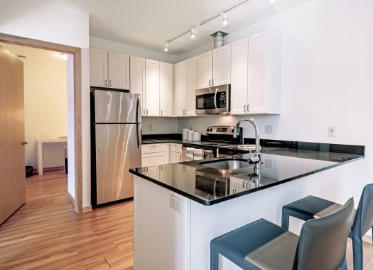 Modern kitchen here at 7west Apartment Homes with white cabinets, stainless steel appliances, black granite countertops, and light wood flooring. Two blue bar stools by the island.
