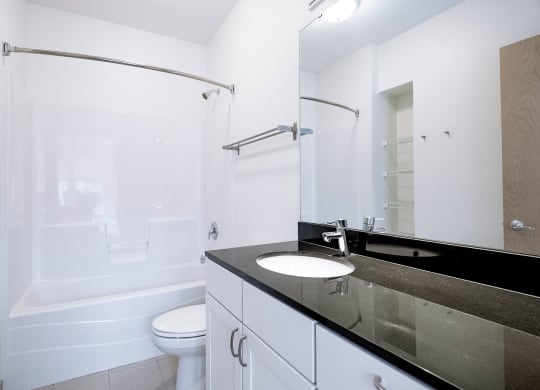 Modern bathroom here at 7west Apartment Homes with white walls and fittings. Features a bathtub with a shower curtain, a toilet, and a sleek black countertop with a sink.