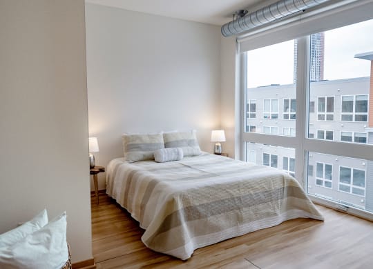 A modern bedroom here at 7west Apartment Homes with a neatly made bed featuring striped bedding. Two lamps on bedside tables provide soft lighting. Large windows reveal a city view.