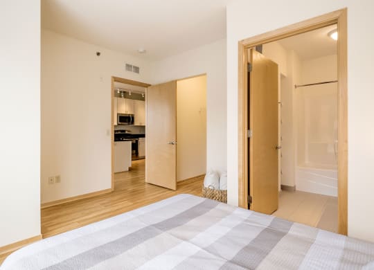 A bright bedroom here at 7west Apartment Homes featuring a neatly made bed, light wood floor, and cream walls. Open doors reveal a modern kitchen and a bathroom, creating a cozy, inviting ambiance.