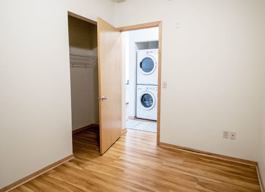 Empty room here at 7west Apartment Homes with light wood flooring, open door leading to a closet, and view of stacked washer and dryer in adjoining room. Bright and minimalistic.