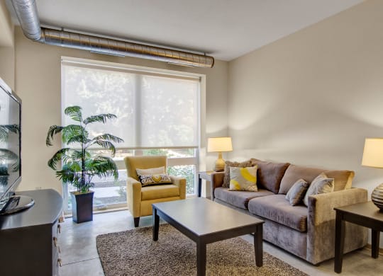 A cozy living room here at 7west Apartment Homes with a brown sofa and yellow armchair next to large windows. Warm lighting from lamps and greenery create a relaxed, inviting atmosphere.