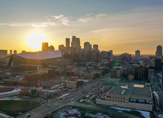 Sunset over a city skyline at 7west Apartment Homes with a prominent, modern stadium. The sun casts a warm glow, highlighting tall buildings and snow-dusted streets below.
