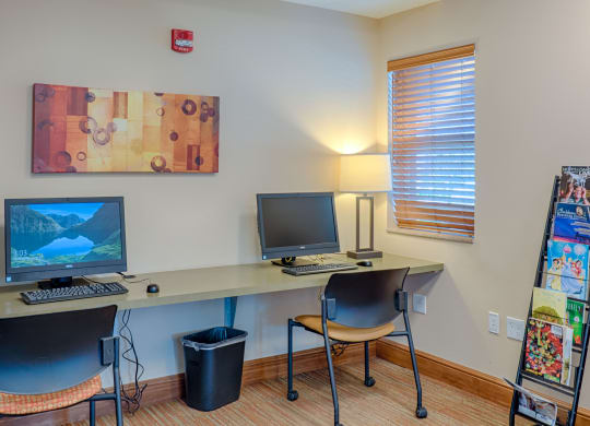 A small office space here at Dakota Ridge Apartment Homes with two computers on a desk, a lamp, and a window with blinds. A magazine rack stands to the right, while a colorful abstract painting adorns the wall, creating a calm and organized atmosphere.