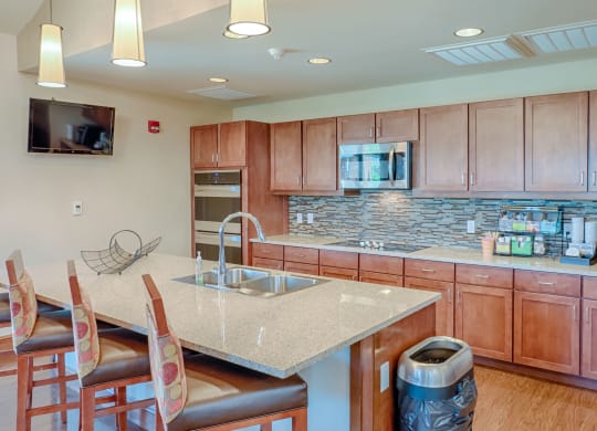 Modern kitchen here at Dakota Ridge Apartment Homes with wooden cabinets, tiled backsplash, and stainless steel appliances. Granite island counter has three chairs and a fruit basket.