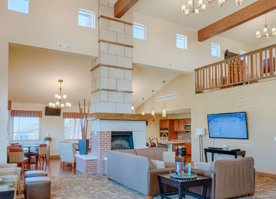 Spacious living room here at Dakota Ridge Apartment Homes with high ceilings, stone fireplace, and wooden beam accents. Cozy seating area, chandelier lighting, and a TV displaying a tennis match.