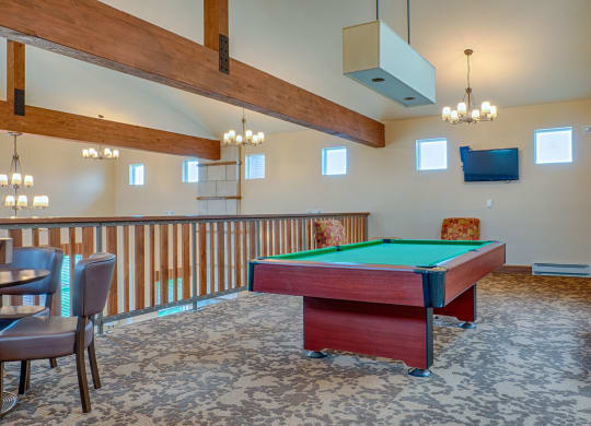 Spacious game room here at Dakota Ridge Apartment Homes with a green-felt pool table under warm lighting. Exposed wooden beams, carpeted floor, TV, and seating area create a cozy ambiance.