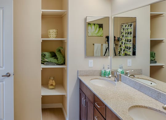 Modern bathroom here at Dakota Ridge Apartment Homes with dual sinks, granite countertop, and mirror. Open shelves hold green towels and decorative items. Bright, tidy, and inviting.