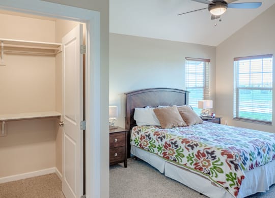 Cozy bedroom here at Dakota Ridge Apartment Homes with a colorful floral bedspread, wooden headboard, and bedside table. Open closet with shelves. Warm light streams through large windows.