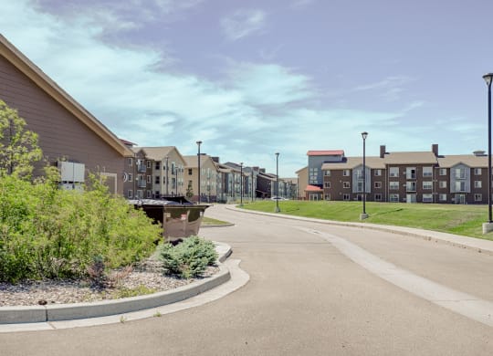 Wide suburban street with modern Dakota Ridge Apartment Homes buildings on the right. Lush greenery and a clear blue sky create a calm, spacious suburban feel.
