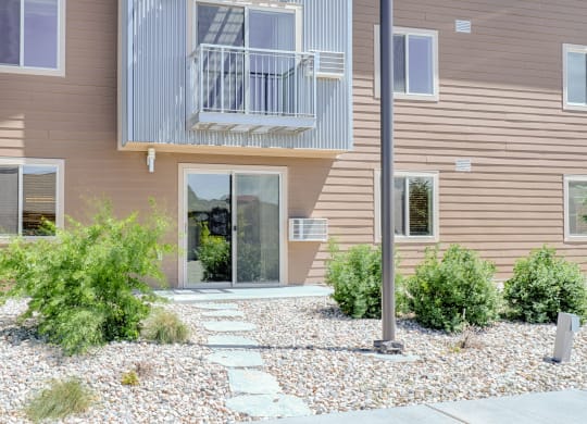 Two-story building exterior here at Dakota Ridge Apartment Homes with brown siding and metal balcony. A path of flat stones leads to a glass door, surrounded by green shrubs and pebbles.