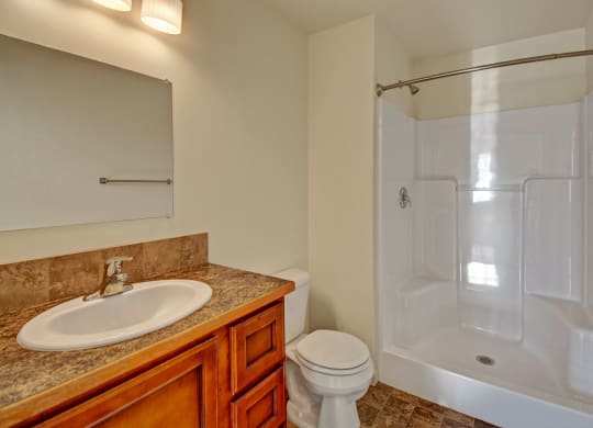 Compact bathroom here at Phoenix Ridge Apartment Homes with a white shower, beige walls, and tiled floor. Features a wooden vanity, brown countertop, sink, and toilet, under warm lighting.