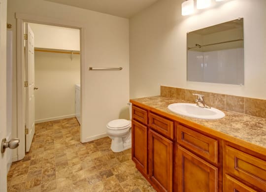 Spacious bathroom here at Phoenix Ridge Apartment Homes with wood vanity, marble countertop, and mirror. Toilet beside a door leading to laundry room. Warm, clean, and functional atmosphere.