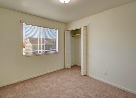 Empty bedroom here at Phoenix Ridge Apartment Homes with beige carpet, cream walls, and a window with blinds. An open closet with a white door is on the right, under a ceiling light.