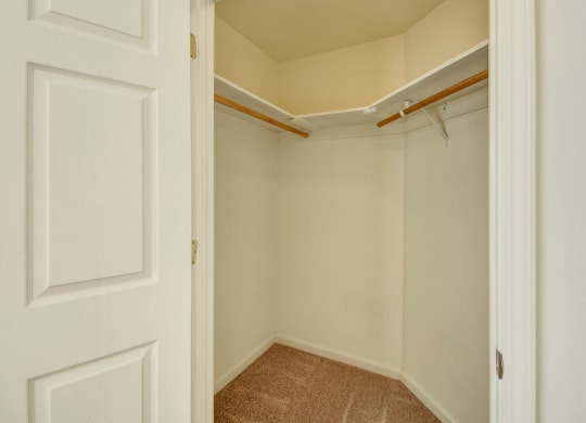 Empty walk-in closet here at Phoenix Ridge Apartment Homes with beige walls, carpeted floor, and wooden hanging rods on white shelves. The open door reveals a spacious, clean feel.