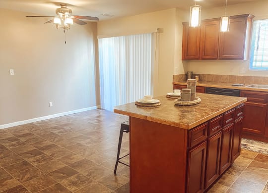 Warm kitchen here at Phoenix Ridge Apartment Homes with brown cabinets and tile floor. An island with dishes and stools. Soft lighting from ceiling fixtures creates a cozy atmosphere.