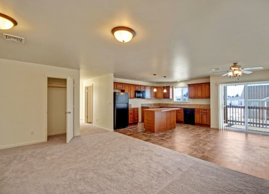 Spacious open-plan living area here at Phoenix Ridge Apartment Homes with beige carpet and tiled kitchen. Features wooden cabinets, center island, glass doors to balcony, and a cozy, bright ambiance.