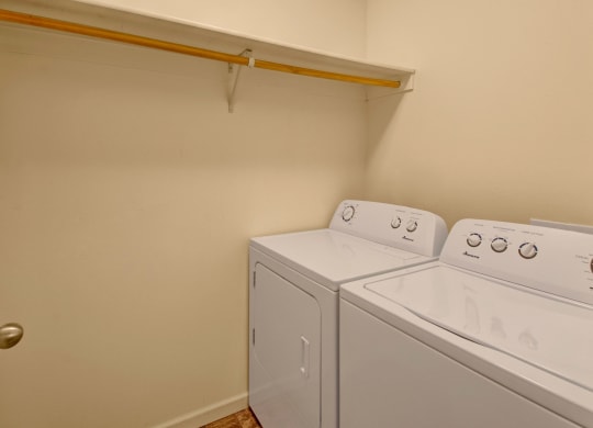 Small laundry room here at Phoenix Ridge Apartment Homes with white washer and dryer against a beige wall. A wooden shelf with a hanging rod is above the appliances. The space is tidy and functional.