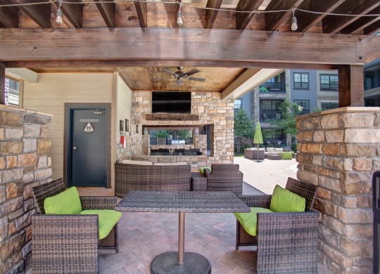 Covered patio area here at the Enclave at Brookside Apartments with wicker furniture and lime green cushions. Stone walls and wooden beams complement a TV above a fireplace. Relaxed, inviting atmosphere.