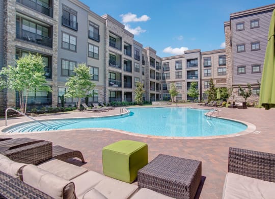 Spacious swimming pool surrounded by the Enclave at Brookside's modern apartment complex under a clear blue sky. Wicker furniture with green cushions adds a cozy touch.