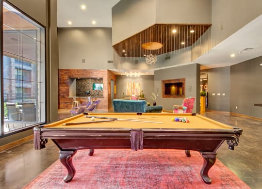 Spacious lounge here at the Enclave at Brookside Apartments with a central pool table on a red rug. Modern decor, high ceilings, large windows, comfortable seating, and a cozy atmosphere.