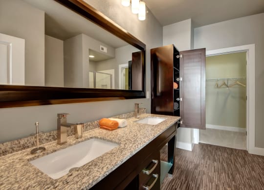 Modern bathroom here at the Enclave at Brookside Apartments with a granite double vanity, sleek faucets, and a large mirror. An open wooden cabinet reveals neatly folded towels. A walk-in closet is visible through an open door.
