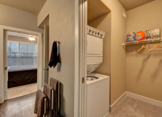 Stacked washer and dryer in a compact laundry area here at the Enclave at Brookside Apartments, beige walls, shelf with detergent, and hangers. Open door reveals bedroom with large window. Cozy ambiance.