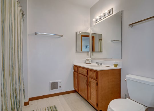 Small bathroom with a white toilet, wooden vanity, and large mirror. Light fixture above, and striped shower curtain to the left. Calm, minimal look. Arbor Ridge Apartments