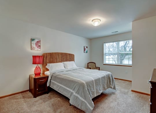 Cozy bedroom at Arbor Ridge Apartments with a woven headboard bed, gray quilt, and red lamp on a wooden nightstand. Soft lighting, window view, and floral art create a calming ambiance.
