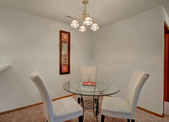 Dining area in Arbor Ridge Apartments featuring a round glass table with three upholstered chairs, a chandelier with five lights above the table, white walls, a decorative mirror on one wall, and light brown carpeted flooring.