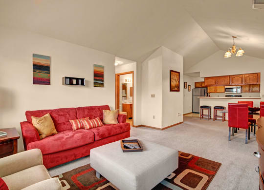 Cozy living room at Arbor Ridge Apartments with a red sofa, beige armchair, and geometric rug. Open dining area features red chairs and wooden kitchen cabinets, creating a warm, inviting atmosphere.