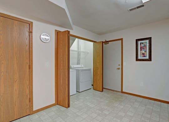 Laundry room with open wooden bi-fold door at Arbor Ridge Apartments revealing a white washer and dryer. A wall clock and framed art add warmth to the neutral-toned space.