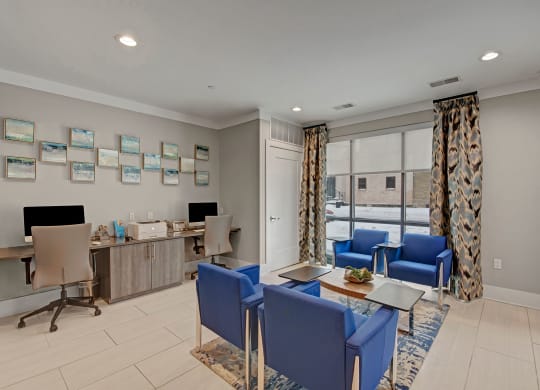 Modern business center with two workstations, computers, and printer on left. Blue chairs around tables in center, large window with patterned curtains behind at River House Apartments.