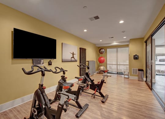 A modern home gym with three stationary bikes, a wall-mounted TV, a yoga poster, gym balls, and large windows, creating a focused, serene atmosphere at River House Apartments.