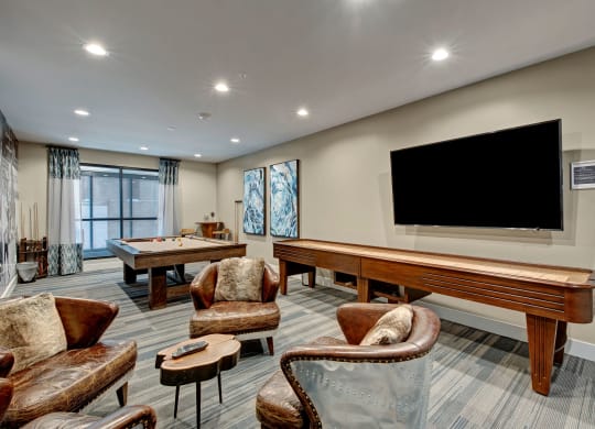 Cozy community room with a pool table, shuffleboard, leather chairs, and a large TV at River House Apartments.