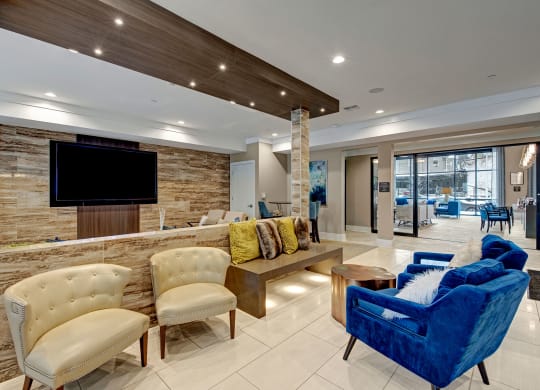 Modern lobby with a sleek, contemporary design. Features cream chairs, a plush blue sofa, colorful cushions, wood accents, and a large TV at River House Apartments.