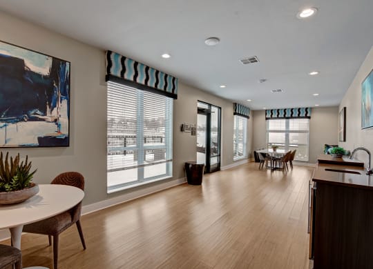 Modern community room with wood flooring and large windows covered by patterned valances. Features abstract art, a round table, chairs, and plants at River House Apartments.