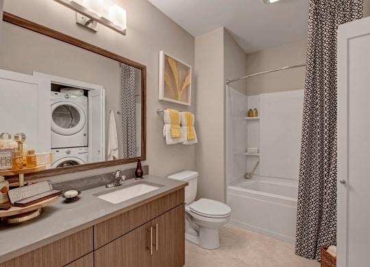 Modern bathroom with wood vanity, large mirror, and stacked washer-dryer. Bathtub with patterned curtain, towels, and framed art create a cozy feel at River House Apartments.