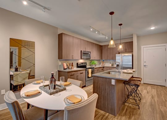Modern kitchen with sleek wood cabinets, stainless steel appliances, island with barstools, and a round dining table set for four. Warm lighting and neutral tones create a cozy atmosphere at River House Apartments.