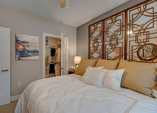 Elegant bedroom with a large, pillowed bed, gold accents, and artistic wall panels. Open closet visible, adding a warm, inviting feel at River House Apartments.