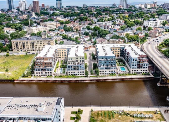 Aerial view of a cityscape featuring River House Apartments along a river. A nearby bridge crosses the water, with a dense urban area in the background.