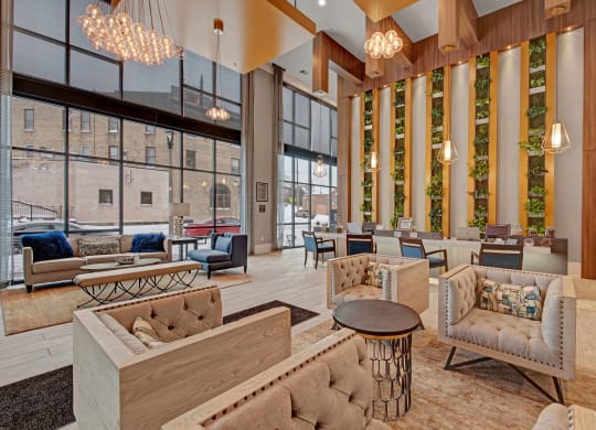 Modern apartment lobby featuring cozy seating with tufted armchairs and sofas, large windows, hanging chandeliers, greenery wall, and a calm ambiance at River House Apartments.