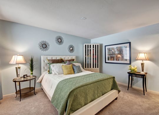 Cozy bedroom with a quilt on a white tufted bed, adorned with colorful pillows. Soft lighting from lamps on side tables creates a serene atmosphere at River's Cove Apartments.