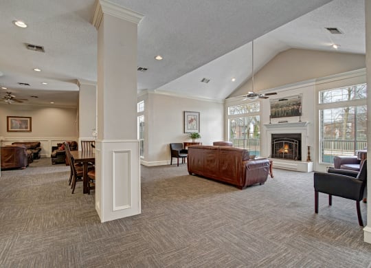 Spacious community room at St. James Place with vaulted ceiling, large windows, and modern gray carpet. Features brown leather sofas, a roaring fireplace, and soft lighting.