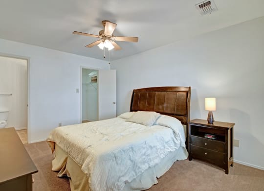 A cozy bedroom featuring a large bed with a wooden headboard and cream bedding. A lit lamp sits on a nightstand, creating a warm atmosphere. Doors lead to a closet and bathroom at St. James Place Apartments.