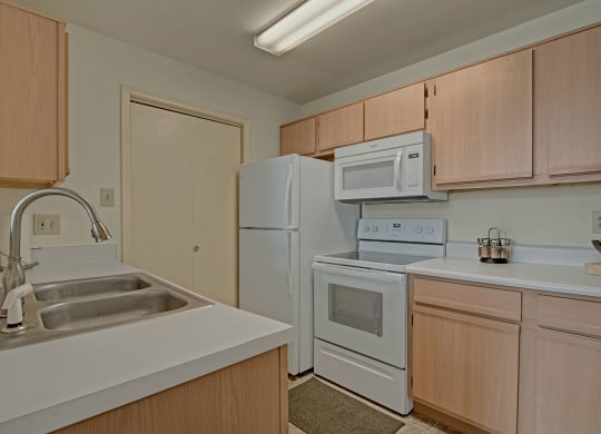 Clean, modern kitchen with light wood cabinets, white appliances, including a stove, microwave, and fridge. Double sink and a bowl of green apples at St. James Place Apartments.