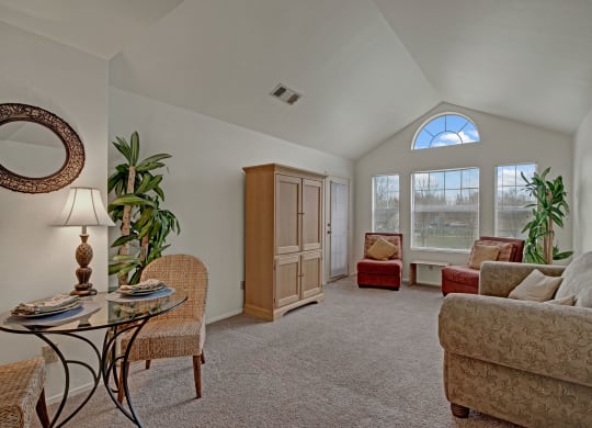 Cozy living room with vaulted ceiling, featuring a beige sofa, two red armchairs, a wooden cabinet, and large windows letting in natural light at St. James Place Apartments.